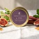 A round tin of Brown Sugar & Fig confectionery sits on a white linen surface with fresh figs, rhubarb, mint, and loose brown sugar scattered around it. The background is a warm neutral wall with soft sunlight and shadows.