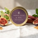 A round tin of Brown Sugar & Fig confectionery sits on a white linen surface with fresh figs, rhubarb, mint, and loose brown sugar scattered around it. The background is a warm neutral wall with soft sunlight and shadows.