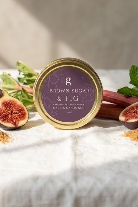 A round tin of Brown Sugar & Fig confectionery sits on a white linen surface with fresh figs, rhubarb, mint, and loose brown sugar scattered around it. The background is a warm neutral wall with soft sunlight and shadows.