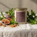 A glass jar candle with a gold lid and a mauve label sits on a white linen tablecloth surrounded by fresh figs, mint leaves, and brown sugar pieces, with warm sunlight casting shadows on a textured beige wall.