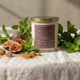 A glass jar candle with a gold lid and a mauve label sits on a white linen tablecloth surrounded by fresh figs, mint leaves, and brown sugar pieces, with warm sunlight casting shadows on a textured beige wall.