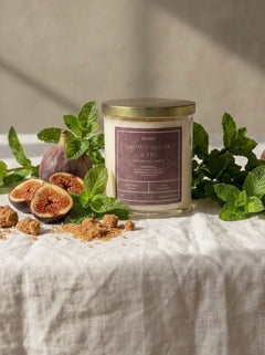 A glass jar candle with a gold lid and a mauve label sits on a white linen tablecloth surrounded by fresh figs, mint leaves, and brown sugar pieces, with warm sunlight casting shadows on a textured beige wall.