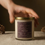 A hand reaches toward a glass jar candle labeled 'Brown Sugar & Co' on a linen-covered table, with dried flowers and driftwood props against a dark brown background.