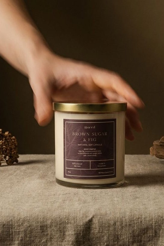A hand reaches toward a glass jar candle labeled 'Brown Sugar & Co' on a linen-covered table, with dried flowers and driftwood props against a dark brown background.