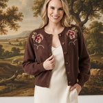 A woman stands indoors wearing a dark brown cropped cardigan with pink floral embroidery over a cream midi dress. The background features a large classical landscape painting and white walls with decorative branches on the floor.