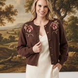 A woman stands indoors wearing a dark brown cropped cardigan with pink floral embroidery over a cream midi dress. The background features a large classical landscape painting and white walls with decorative branches on the floor.