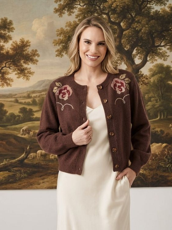 A woman stands indoors wearing a dark brown cropped cardigan with pink floral embroidery over a cream midi dress. The background features a large classical landscape painting and white walls with decorative branches on the floor.