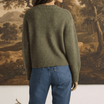 A woman wearing a graced chunky olive green crewneck sweater with a textured knit, paired with blue denim jeans.