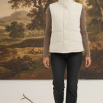 A woman wearing a cream-colored graced corduroy puffer vest over a brown long-sleeve top and dark jeans.