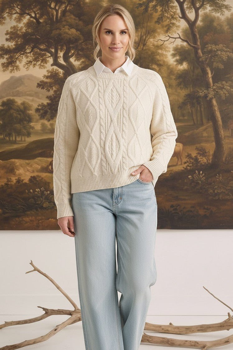 A woman wearing a cream graced crew neck cable knit sweater over a white collared shirt and light wash jeans.
