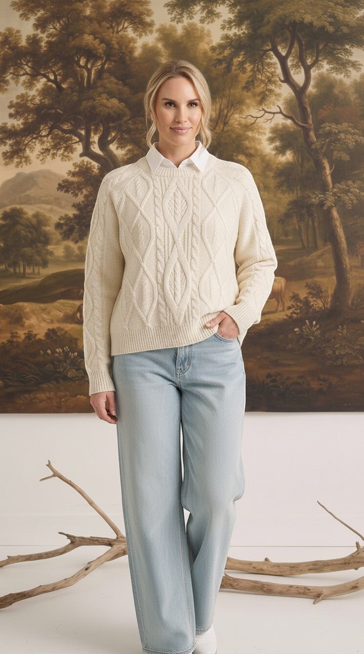 A woman wearing a cream graced crew neck cable knit sweater over a white collared shirt and light wash jeans.