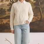 A woman wearing a cream graced crew neck cable knit sweater over a white collared shirt and light wash jeans.