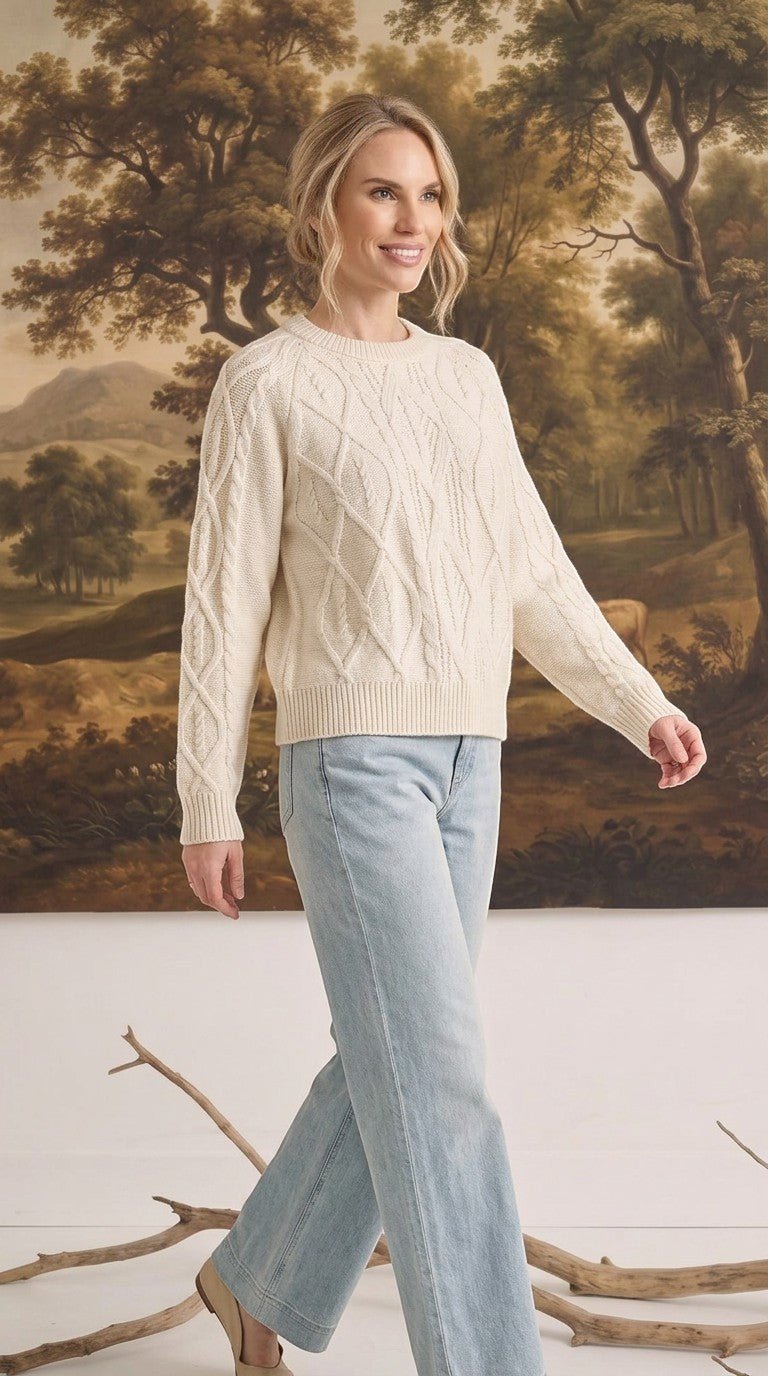A model wearing a cream graced crew neck cable knit sweater with light wash jeans in a studio setting.