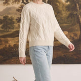 A model wearing a cream graced crew neck cable knit sweater with light wash jeans in a studio setting.