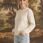 Woman wearing a cream graced crew neck cable knit sweater with light wash jeans in front of a landscape painting