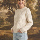 Woman wearing a cream graced crew neck cable knit sweater with light wash jeans in front of a landscape painting