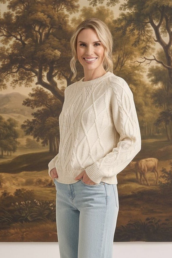 Woman wearing a cream graced crew neck cable knit sweater with light wash jeans in front of a landscape painting