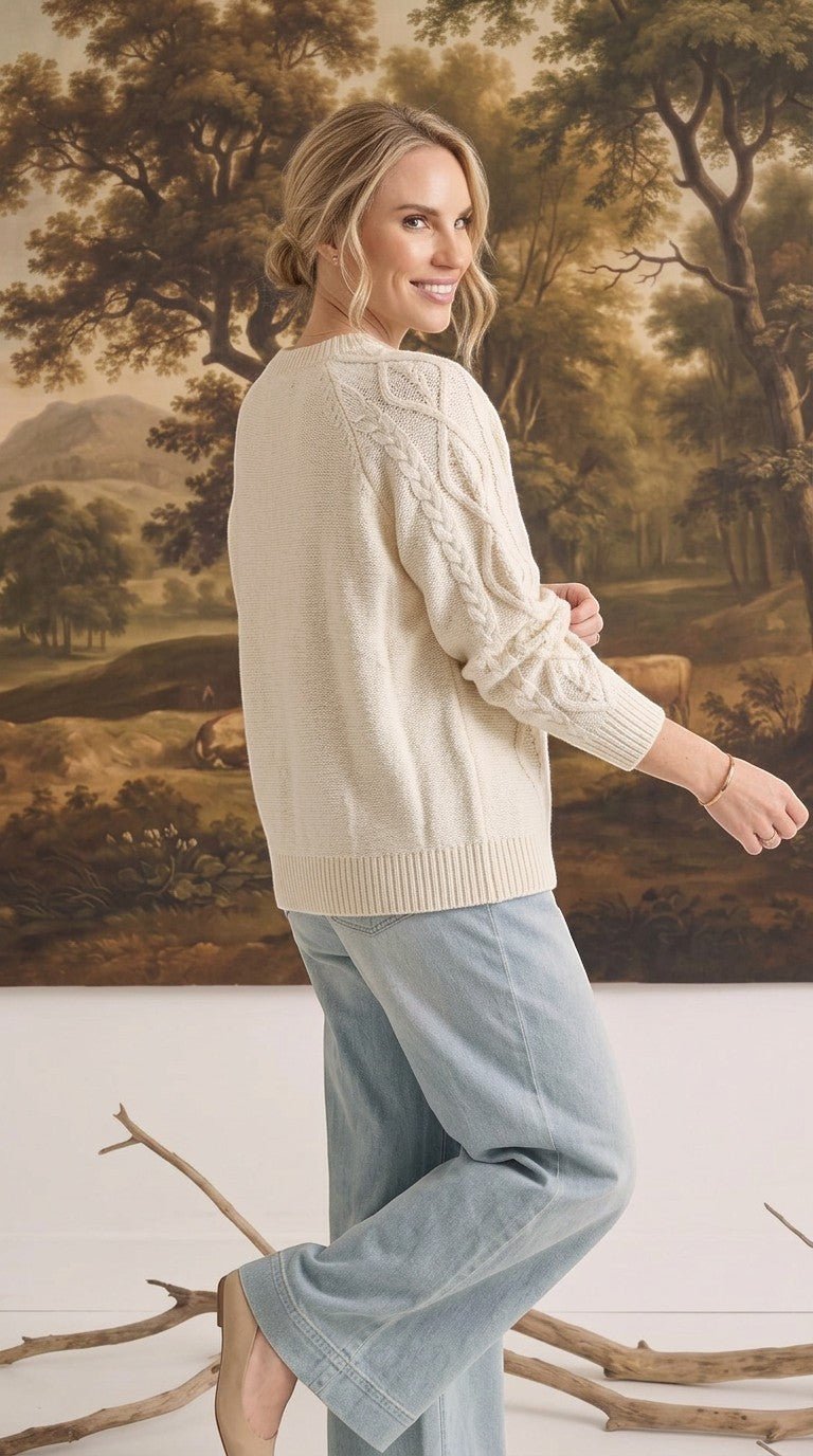 Woman wearing a cream crew neck cable knit sweater by graced, paired with light wash jeans in a studio setting.