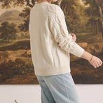 Woman wearing a cream crew neck cable knit sweater by graced, paired with light wash jeans in a studio setting.