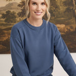 A woman wearing a graced indigo crewneck pullover with ribbed trim, styled with jeans in a studio setting.