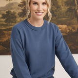 A woman wearing a graced indigo crewneck pullover with ribbed trim, styled with jeans in a studio setting.