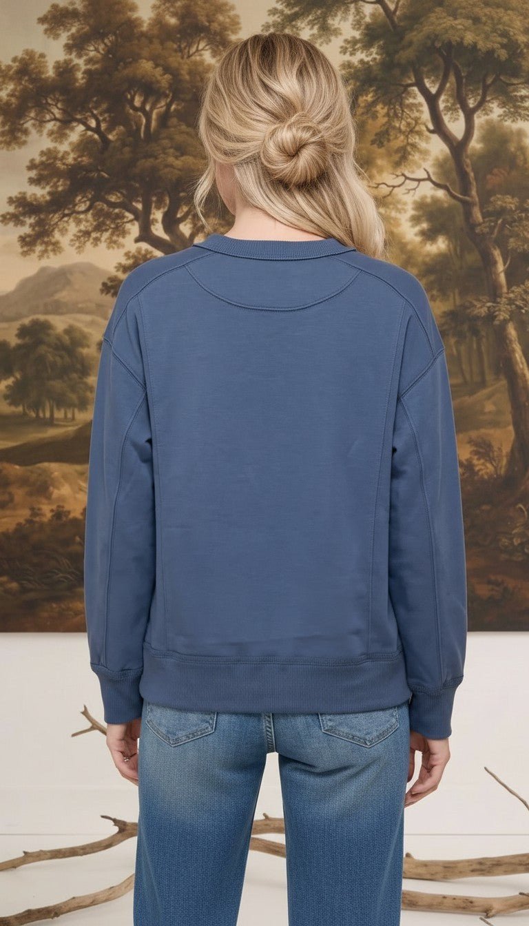 Back view of a woman wearing a graced indigo crewneck pullover with ribbed cuffs and hem against a landscape backdrop