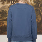 Back view of a woman wearing a graced indigo crewneck pullover with ribbed cuffs and hem against a landscape backdrop