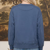 Back view of a woman wearing a graced indigo crewneck pullover with ribbed cuffs and hem against a landscape backdrop