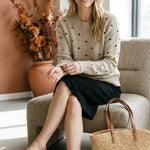 graced beige crewneck sweater with black polka dots worn by a smiling woman sitting in a chair with a straw tote bag