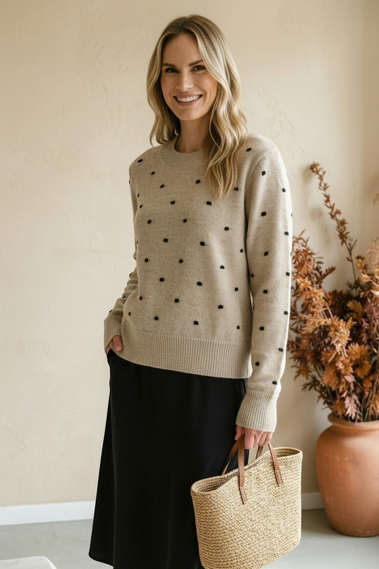 Woman wearing a graced beige crewneck sweater with black polka dots, paired with a black skirt and straw tote bag.