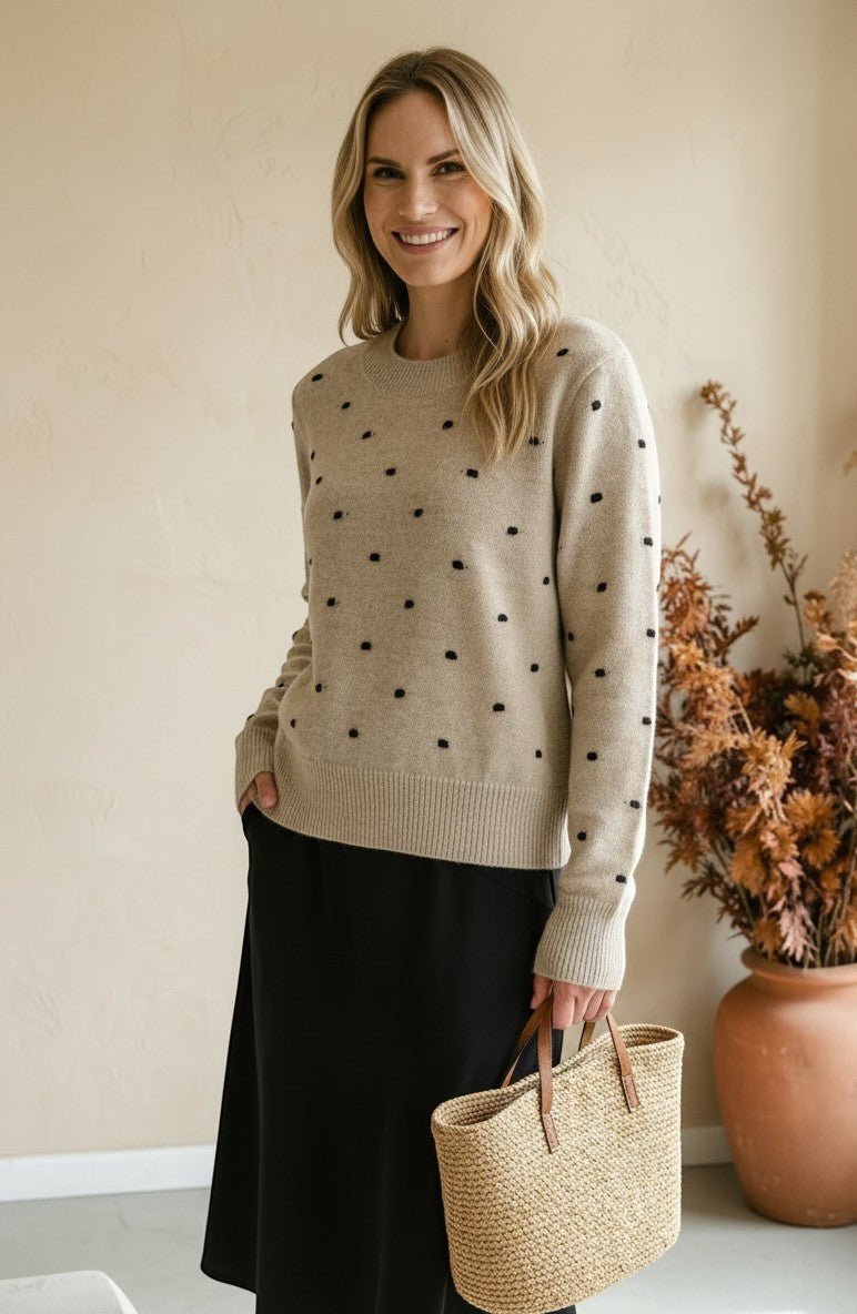 Woman wearing a graced beige crewneck sweater with black polka dots, paired with a black skirt and straw tote bag.