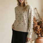 Woman wearing a graced beige crewneck sweater with black polka dots, paired with a black skirt and straw tote bag.