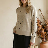 Woman wearing a graced beige crewneck sweater with black polka dots, paired with a black skirt and straw tote bag.