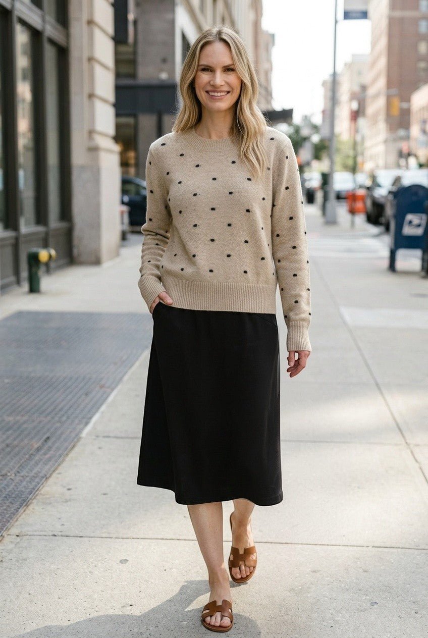 graced beige crewneck sweater with black polka dots worn by a woman with a black midi skirt on a city sidewalk