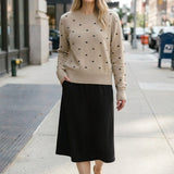 graced beige crewneck sweater with black polka dots worn by a woman with a black midi skirt on a city sidewalk