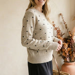 Woman wearing a graced beige crewneck sweater with black polka dots, styled with a black skirt and straw tote bag.