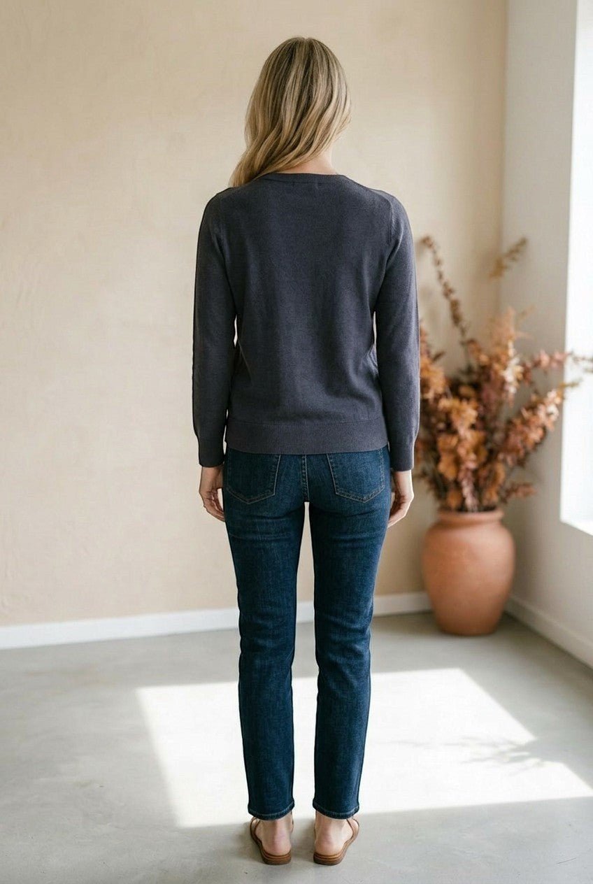 A woman wearing a graced charcoal grey double layered sweater with jeans, viewed from the back in a minimalist room.