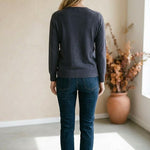 A woman wearing a graced charcoal grey double layered sweater with jeans, viewed from the back in a minimalist room.