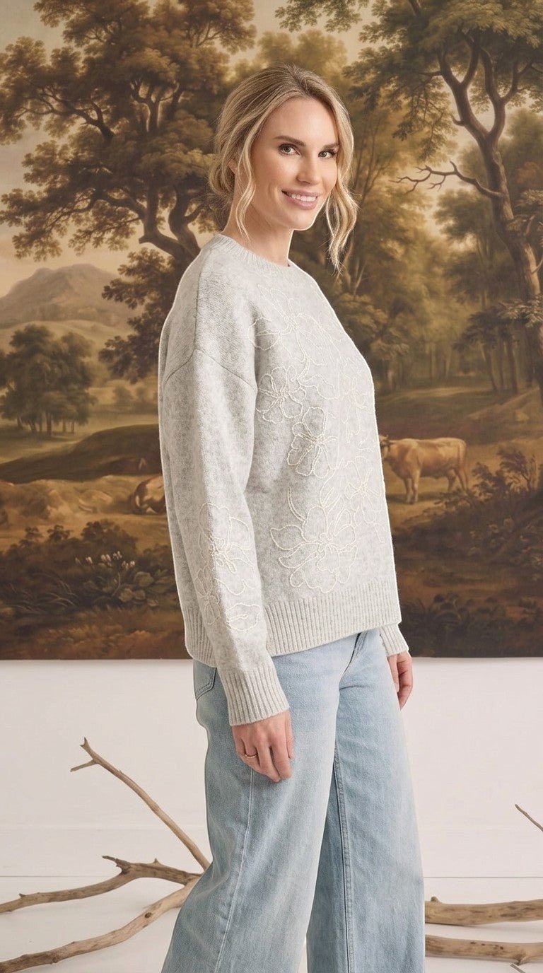 A woman wearing a graced light grey pullover sweater with delicate white floral embroidery on the sleeve and front.