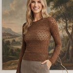A woman modeling a brown textured knit long-sleeve top with a honeycomb pattern, paired with a brown and beige plaid skirt, standing in front of a large classical landscape painting.