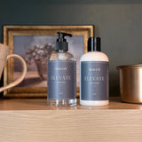 Two bottles of 'Elevate' hand soap and lotion on a wooden surface with a decorative background.