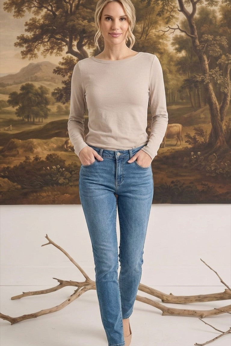 A woman wearing a graced beige long sleeve boat neck knit top tucked into blue jeans against a landscape backdrop.