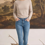 A woman wearing a graced beige long sleeve boat neck knit top tucked into blue jeans against a landscape backdrop.