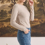 A woman wearing a graced beige long sleeve boat neck knit top tucked into blue jeans against a landscape painting.
