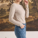 A woman wearing a graced beige long sleeve boat neck knit top tucked into blue jeans against a landscape painting.