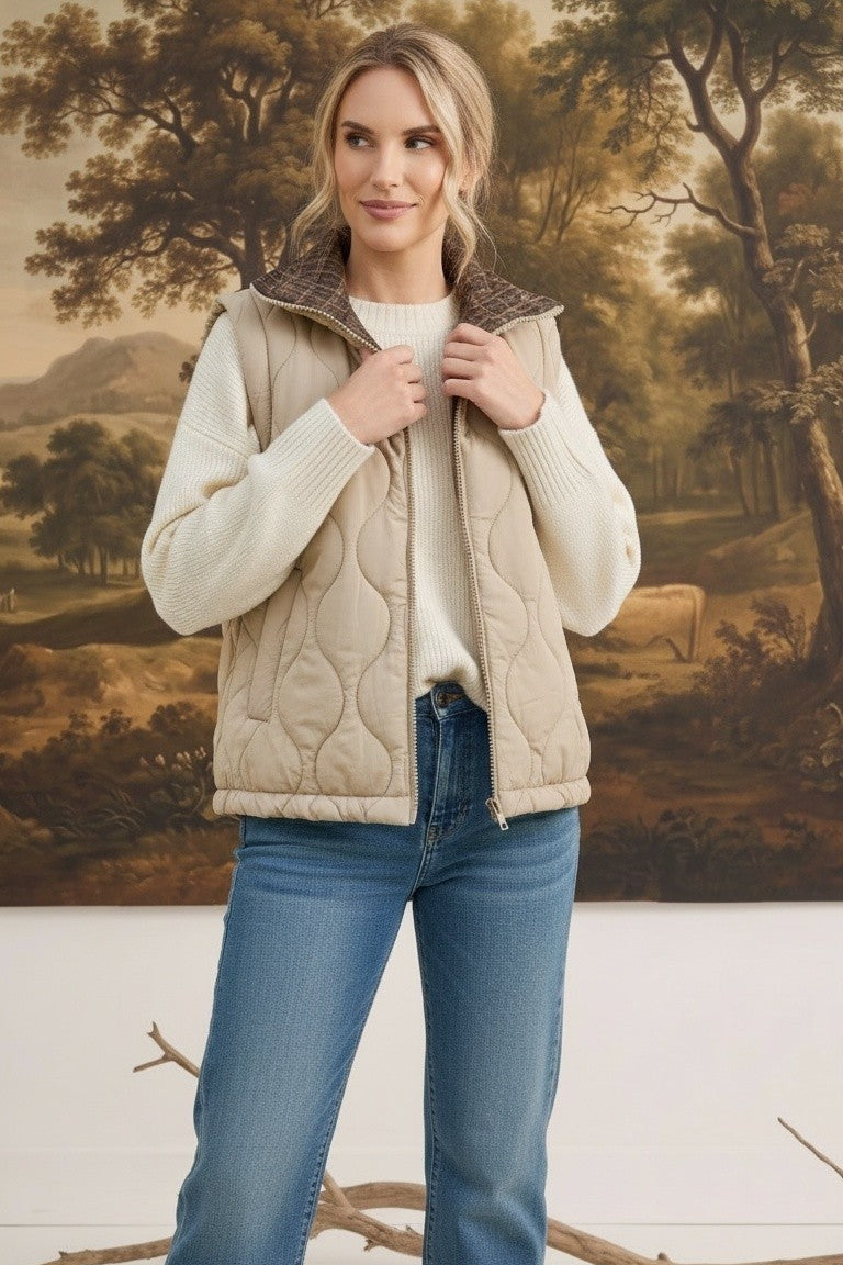 A model wearing a graced beige quilted vest with a plaid collar over a white sweater and blue jeans.