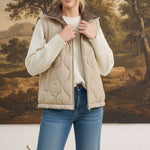 A model wearing a graced beige quilted vest with a plaid collar over a white sweater and blue jeans.
