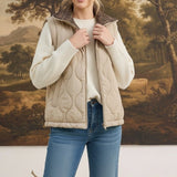 A model wearing a graced beige quilted vest with a plaid collar over a white sweater and blue jeans.