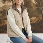 A woman wearing a graced beige quilted vest with a plaid collar over a white sweater and blue jeans.