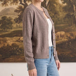 Woman wearing a graced brown crew neck button down cardigan over a white top and blue jeans in a studio setting